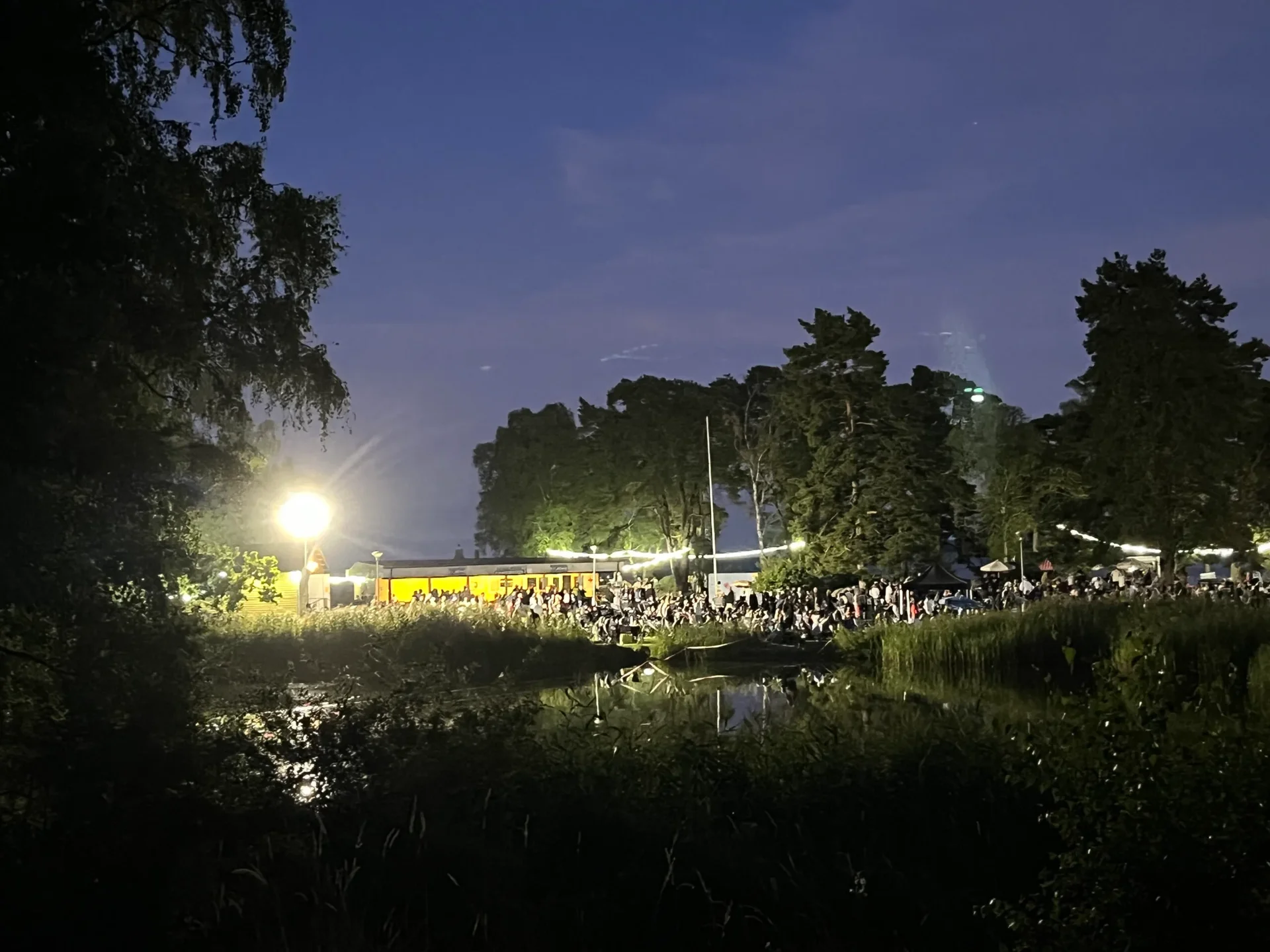 Folksamling vid upplyst byggnad i skymning speglad i vattnet