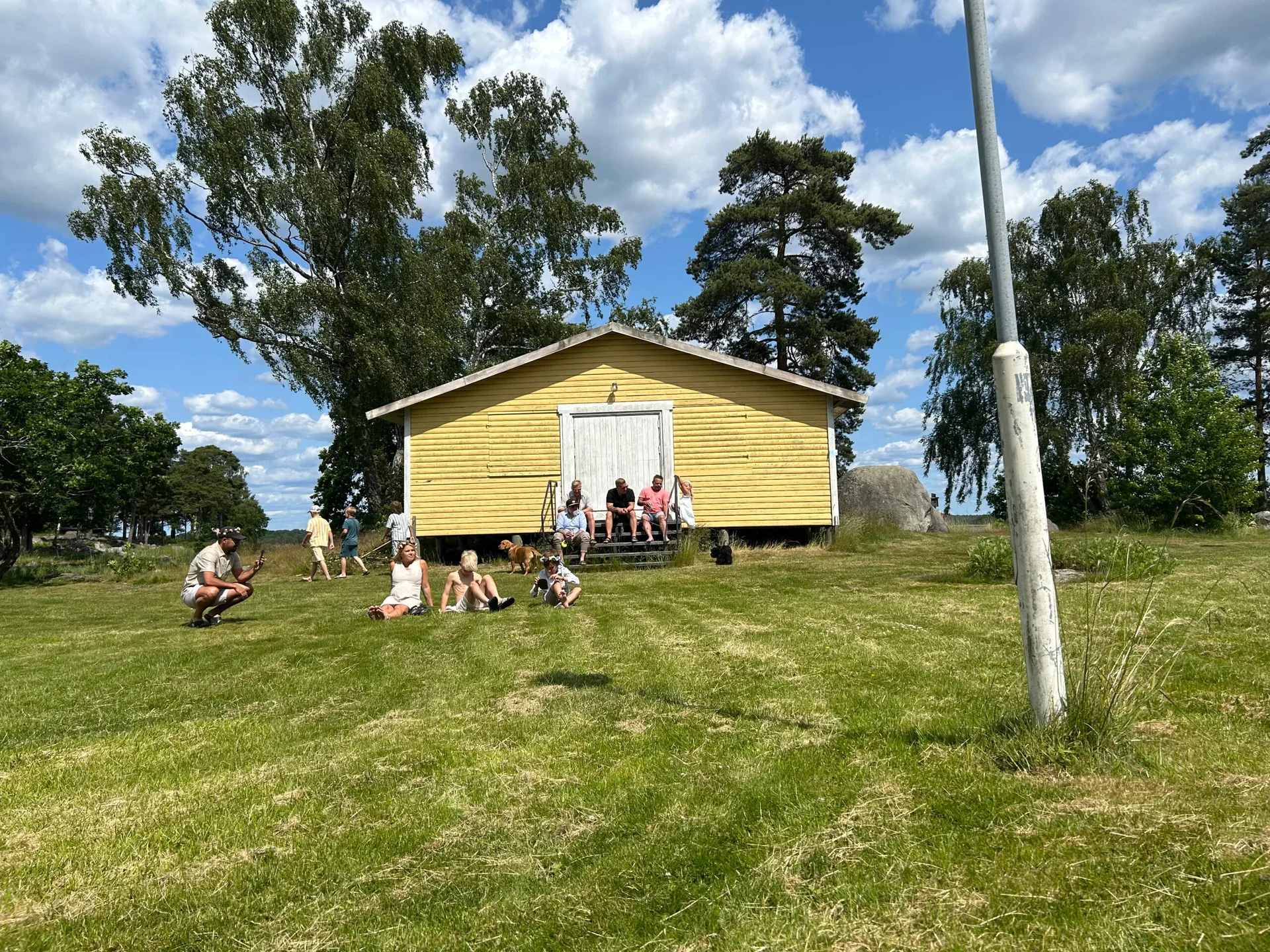 Gul samlingsbyggnad med människor sittande på gräset i solsken
