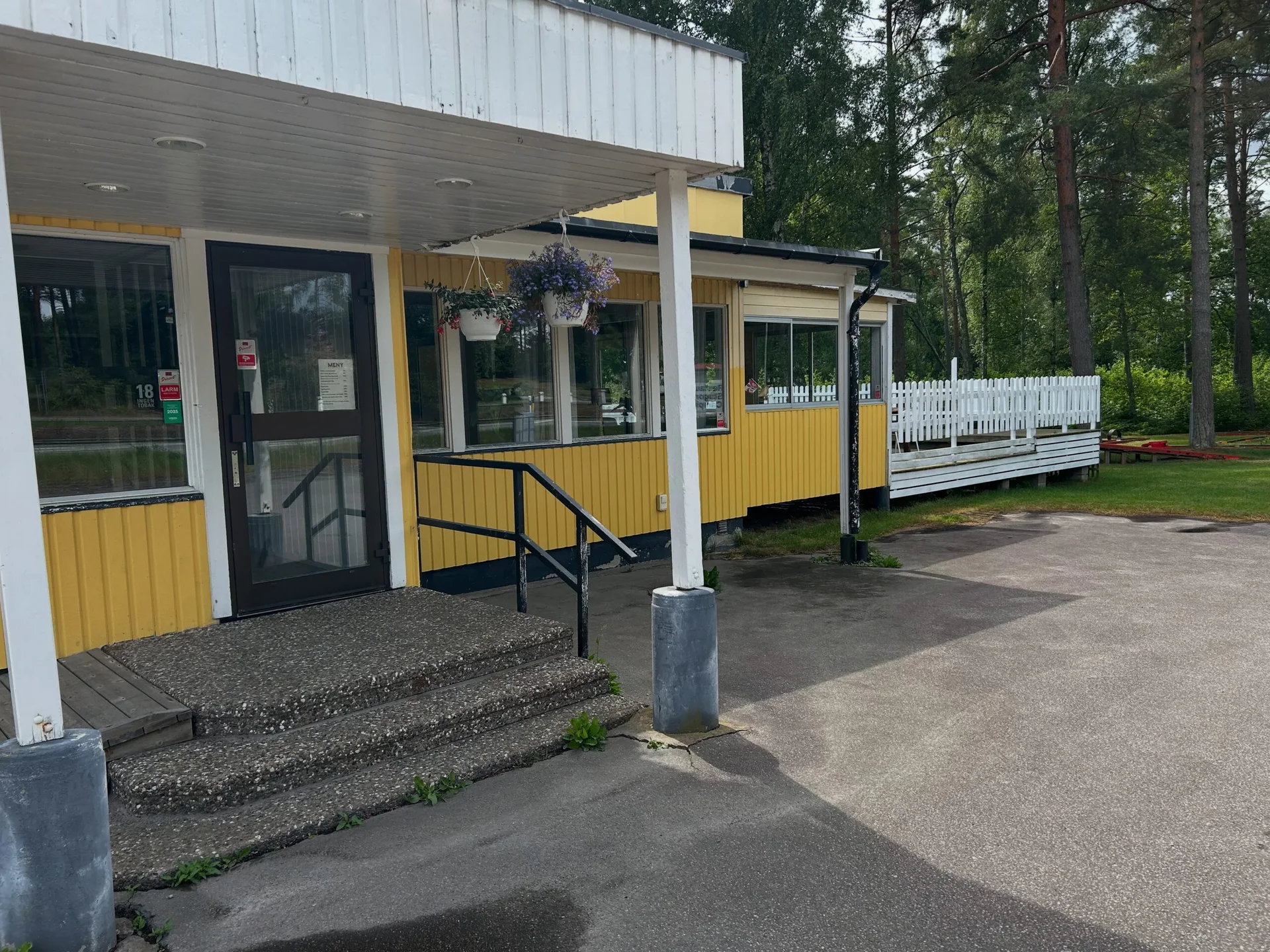 Träspång över vass mot gräsäng och tallar vid vattnet en solig dag